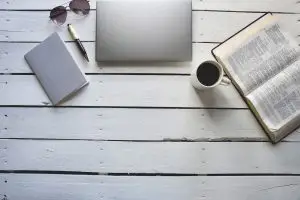 Overhead shot of an open bible near a coffee and, laptob, notepad with a pen
